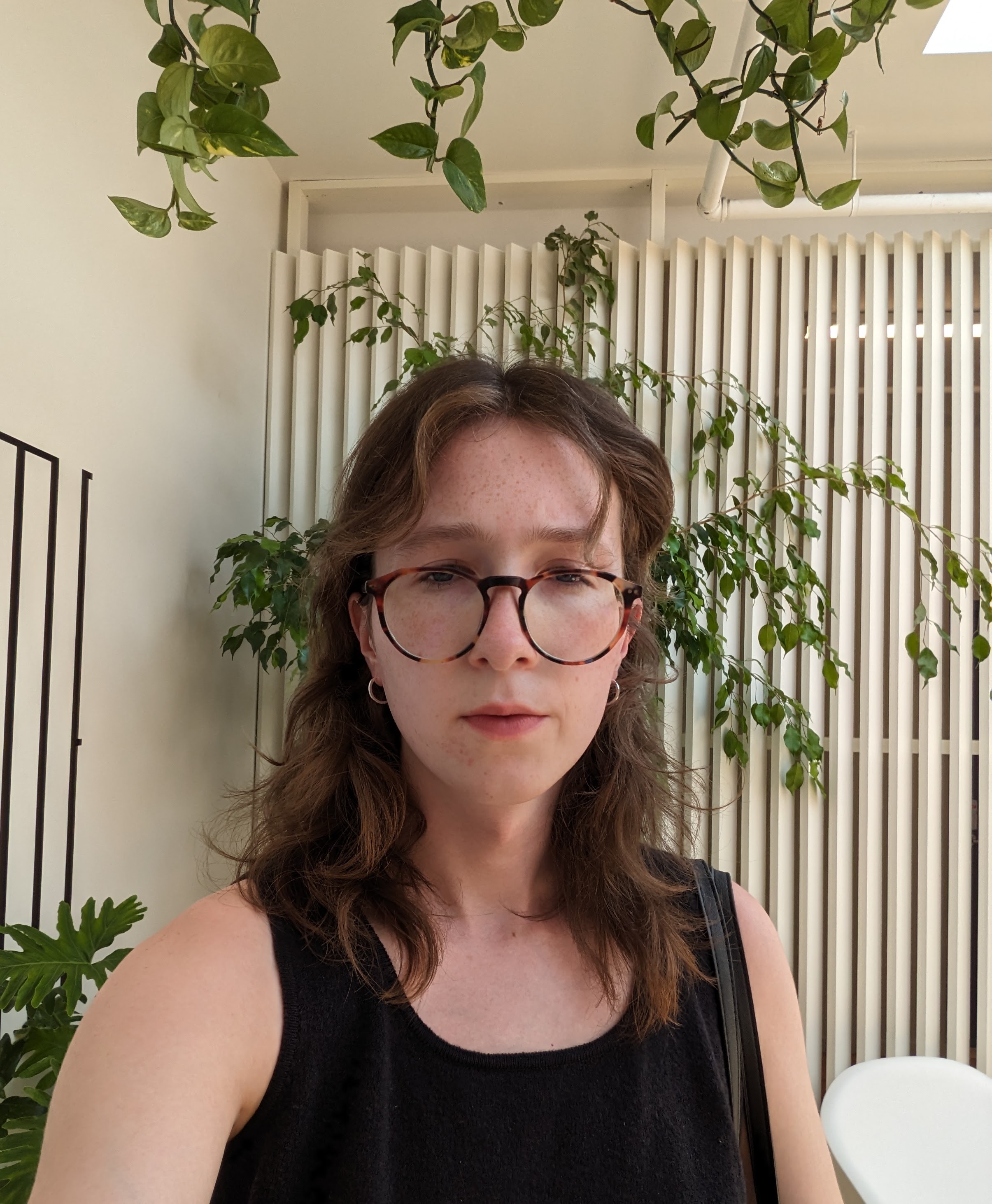 person with light skin tone, shoulder-length curly hair, and glasses in a room with plants