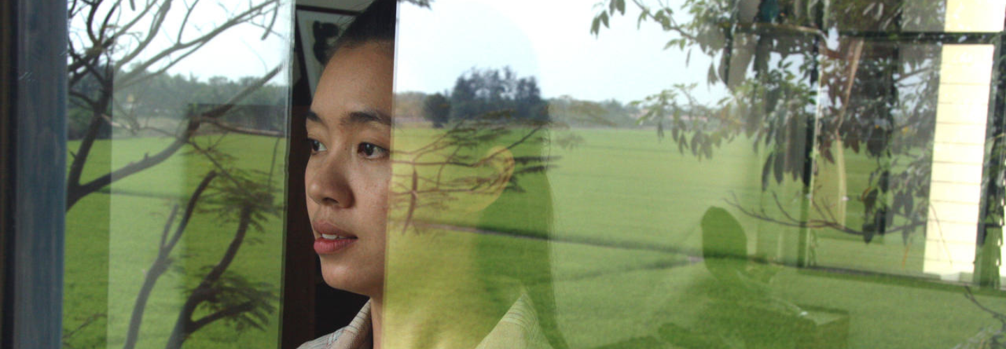 A woman looks out from behind a window, which reflects a green field. 