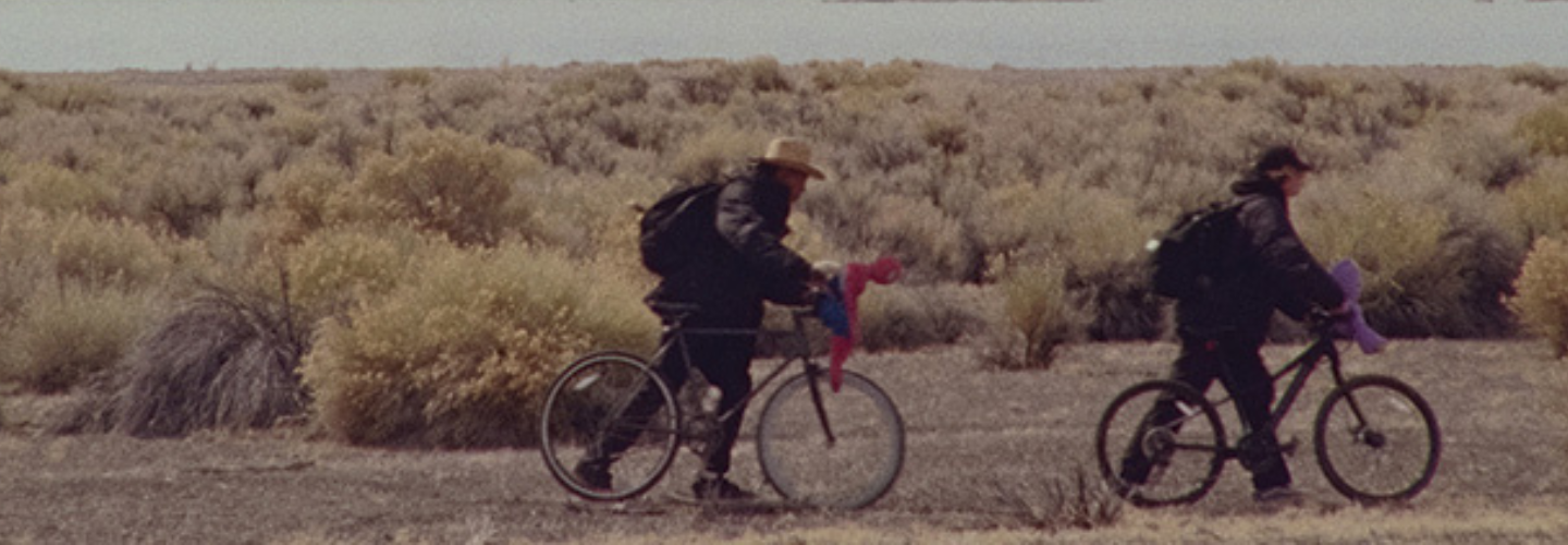 Two people walk with bicycles, a river and snowy mountains in the background