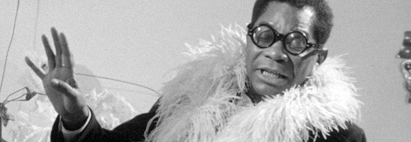 black-and-white film still of man with glasses and feather gesturing with right hand