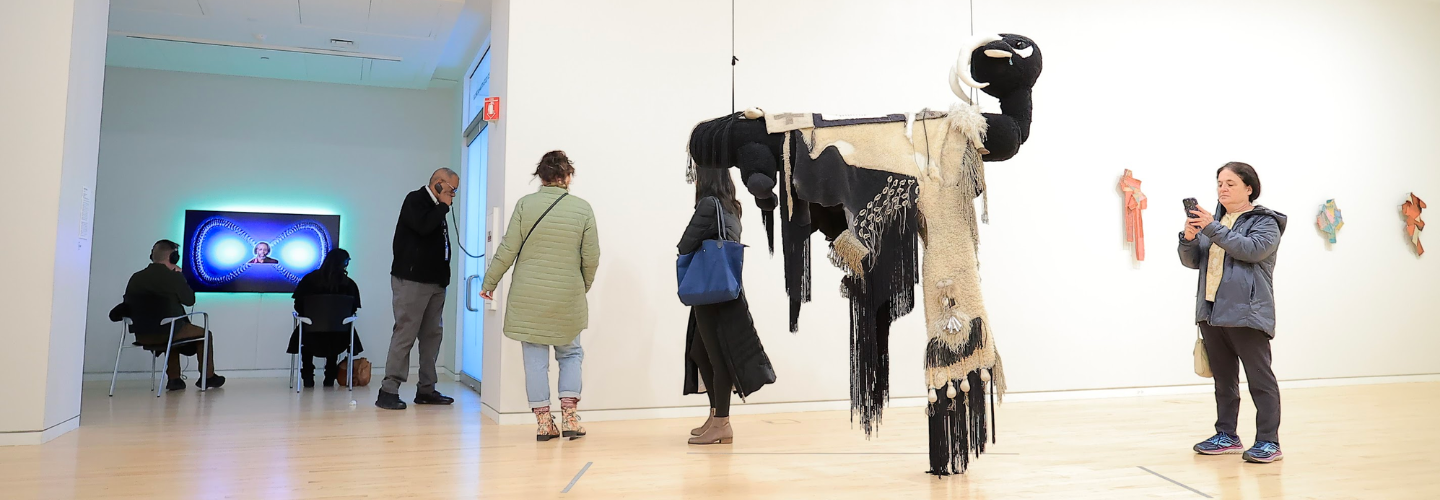 large fiber sculpture of a sheep covered in textiles, hanging from the ceiling in an art gallery with people around