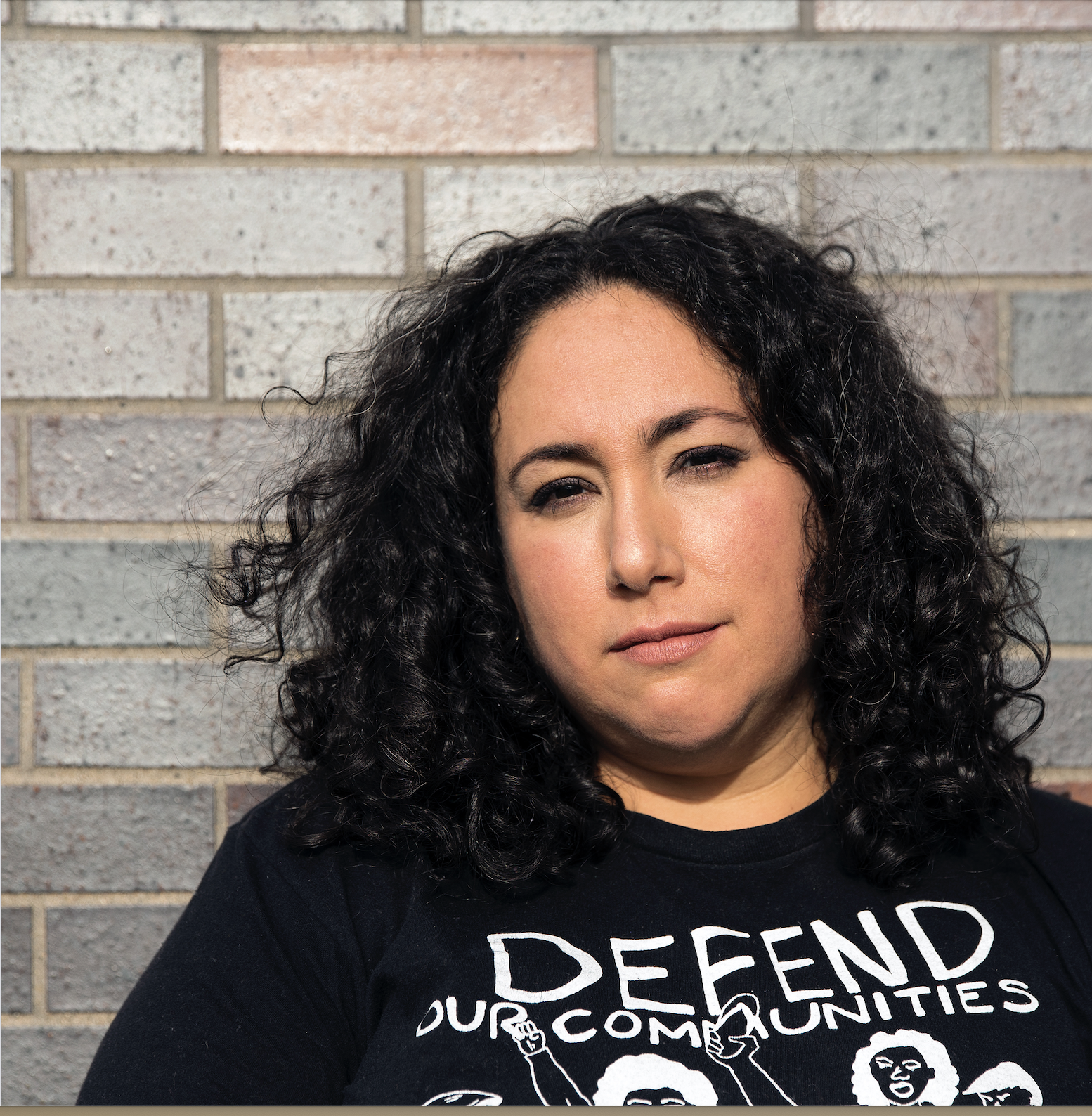 headshot of person with light skin tone and dark curly hair, wearing a shirt that says "defend our communities"