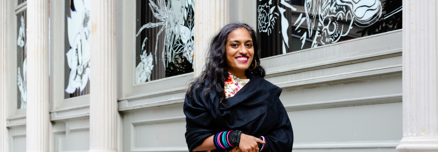 photograph an Indian woman smiling outside a building with artwork on the walls