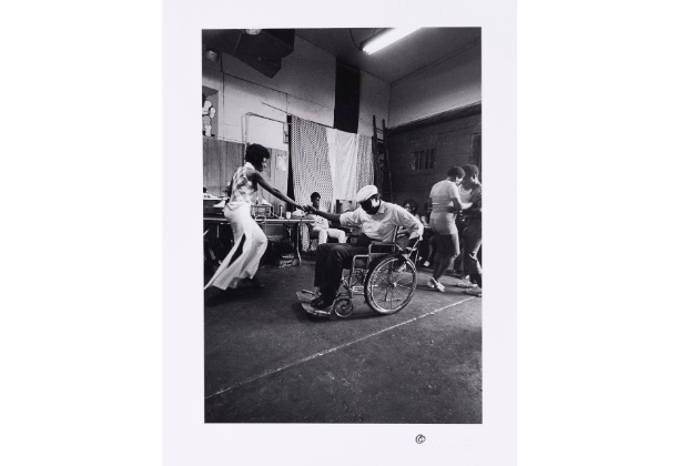 A Black man in a wheelchair smiles and dances with a standing Black woman wearing white bellbottoms