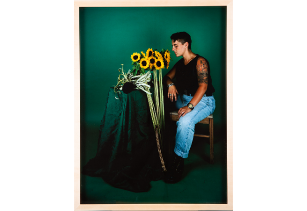 Seated, young, light-skinned adult with tattooed arm faces standing sunflowers in a green room
