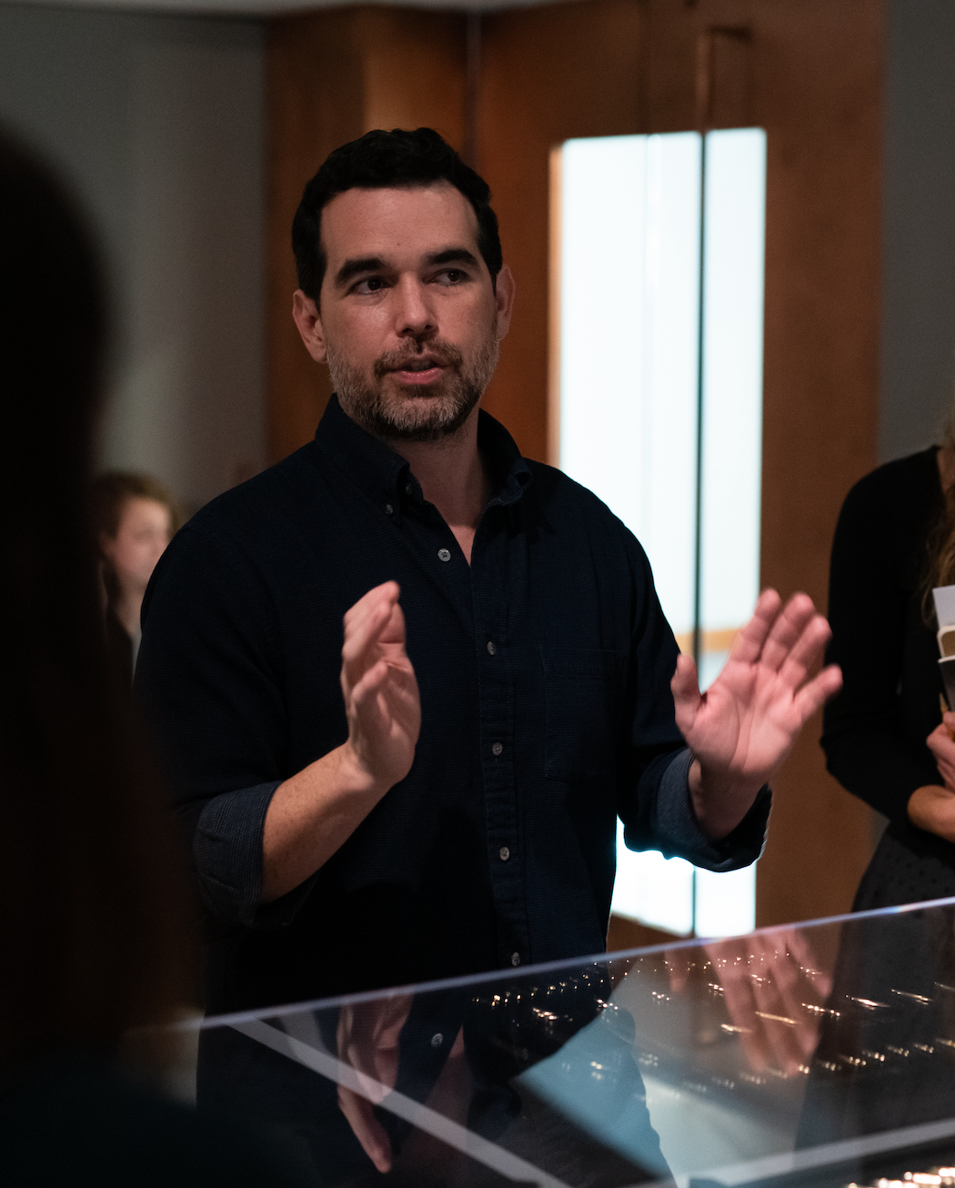 man in front of a glass case, arms raised, as if speaking to a group of people