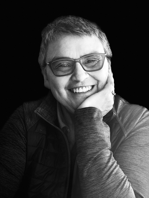 black and white portrait of short-haired woman in glasses, smiling with chin propped up by hand