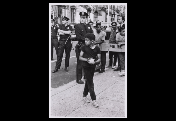 young Black boy holding small ball stands in front of group of police officers