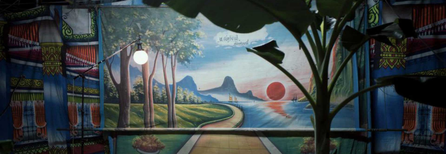 Painted theater backdrops of a rural Thai landscape and setting sun are illuminated by a globe light