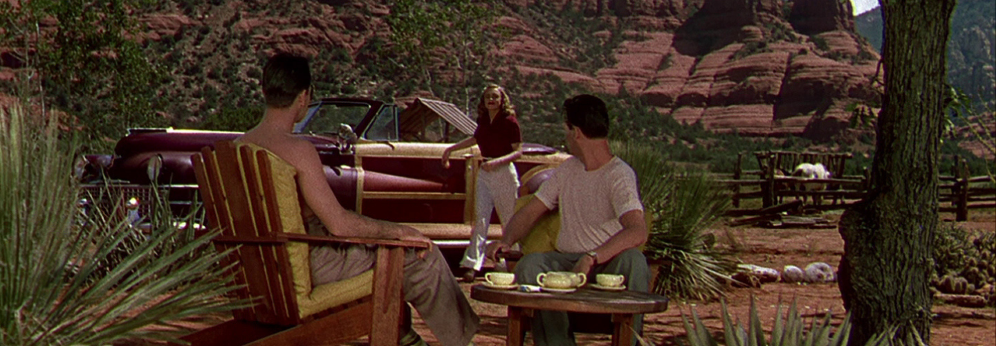 In the center of a vivid desert landscape, two men sit at a table, looking at a woman approach from a 1940s car.