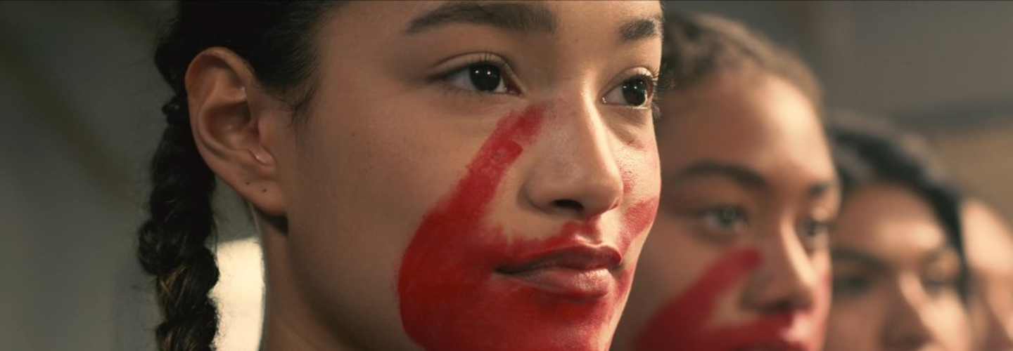 A young woman with brown braided hair, medium skin tone, and red paint on her face.