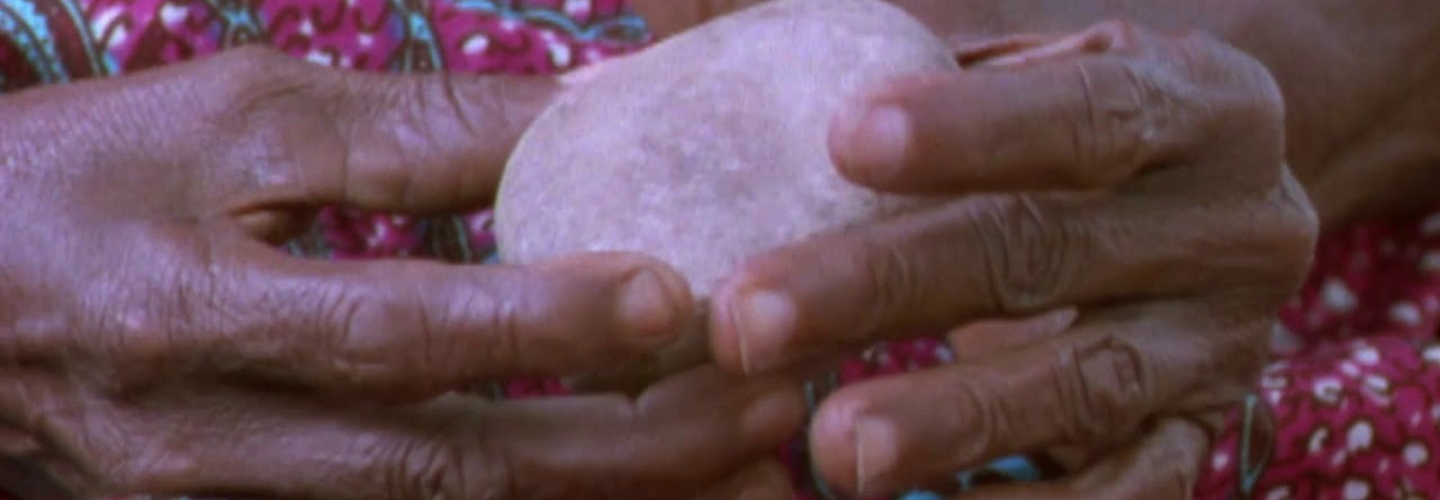 Light pink quartz stone held by hands with dark skin tone. Fuschia and blue printed cloth in the backdrop.