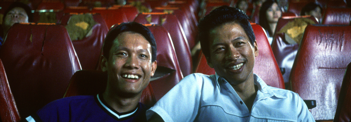 Two young men with medium skin tone sit side to side smiling in a movie theater