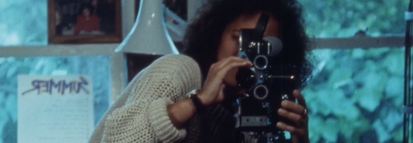 A woman with dark curly hair stands behind a 16mm camera taking her own image