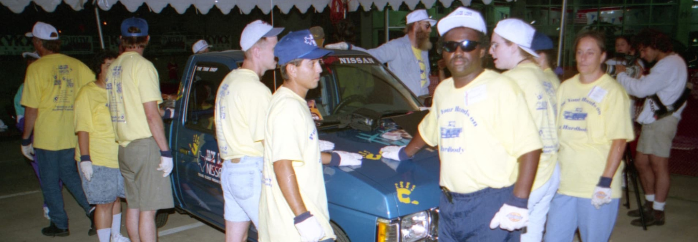 A group of people in yellow shirts and white gloves surround a blue Nissan truck, keeping one hand on it.
