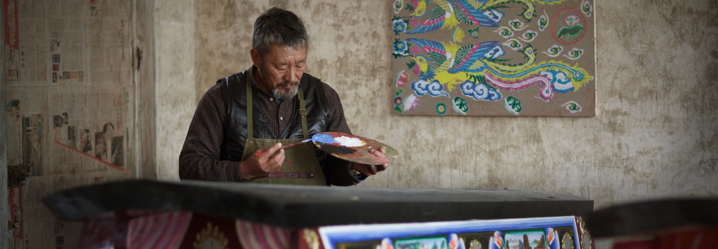 An older man in an arts studio paints a coffin with bright colors 