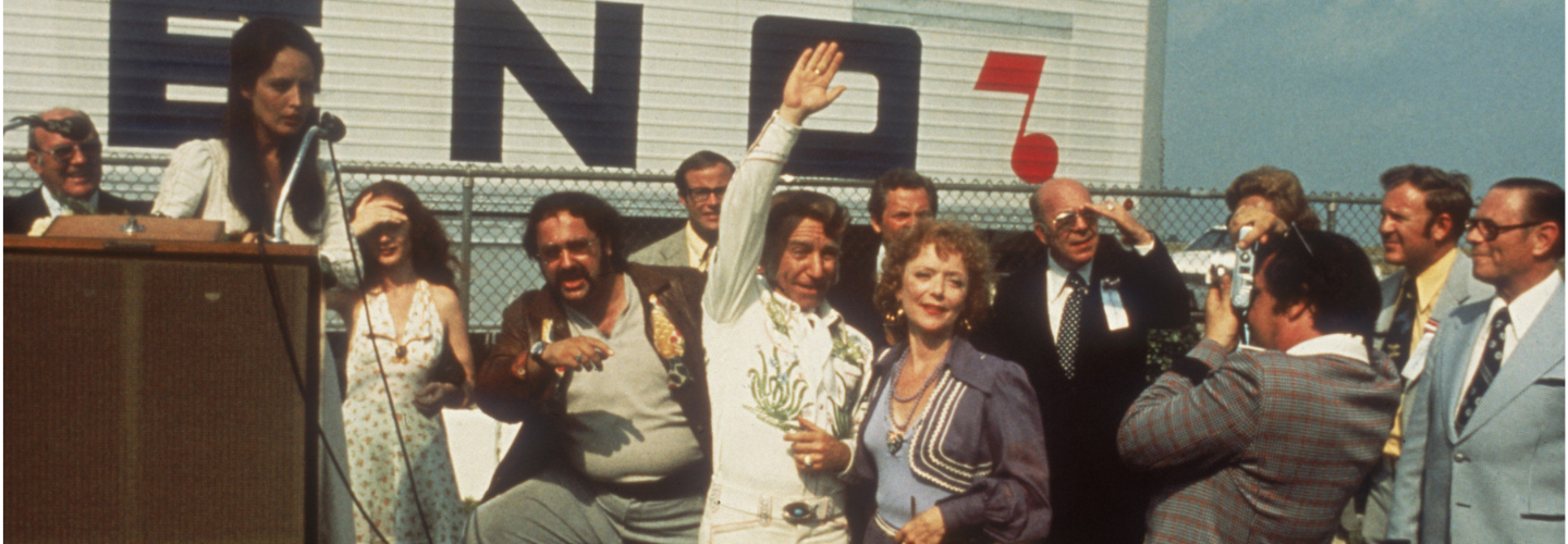 A couple in 70s attire wave to onlookers before stepping onto a stage 