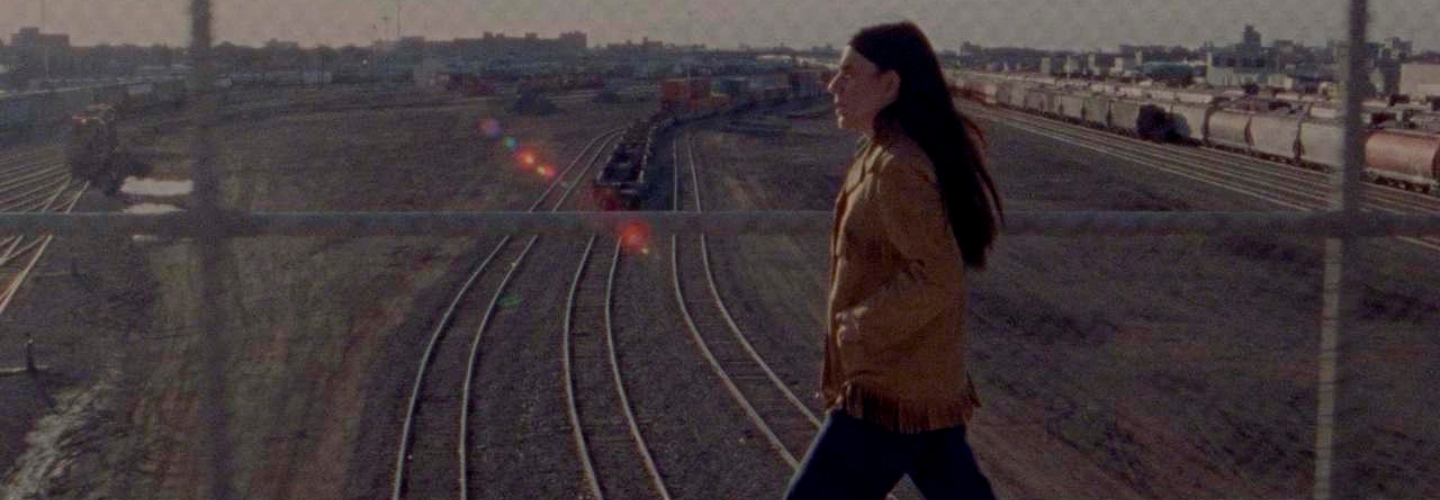 A woman with long dark hair and a tan leather jacket strides across an overpass with train tracks in the background