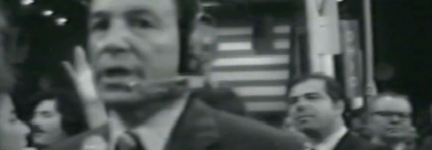 Black and white video image of a newscaster in a suit looking at the camera