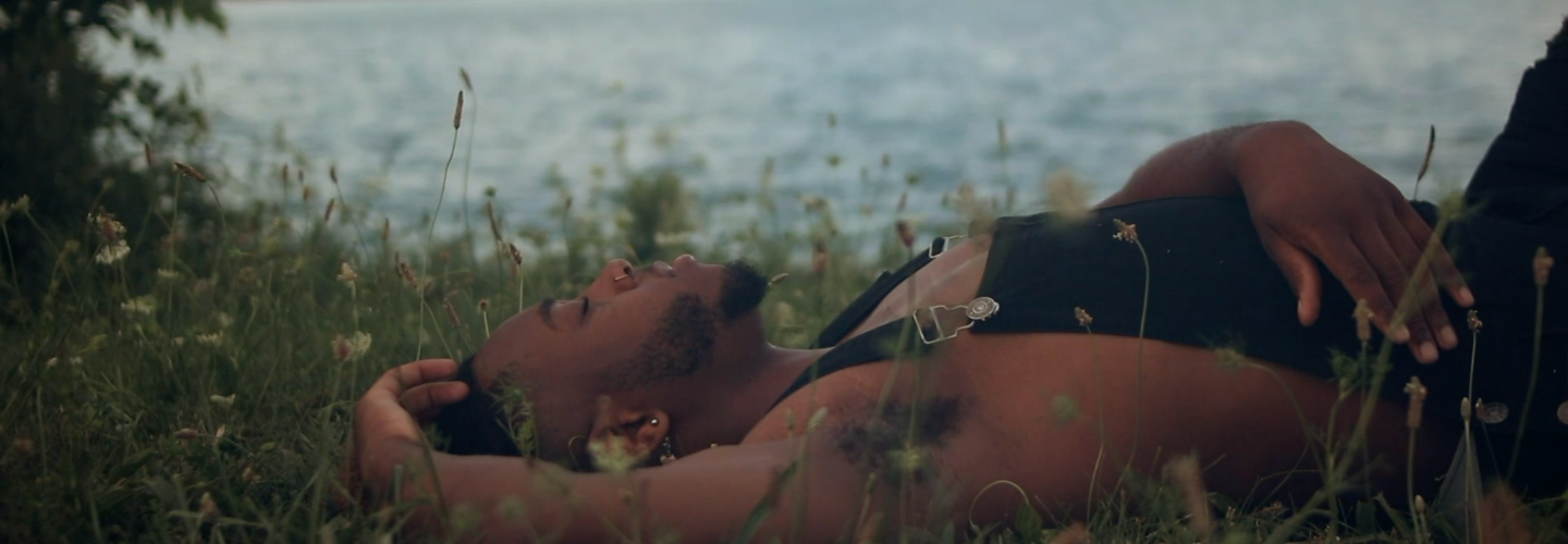Still from NILE. A person with dark skin tone reclining in the grass alongside a lake with a city in the background