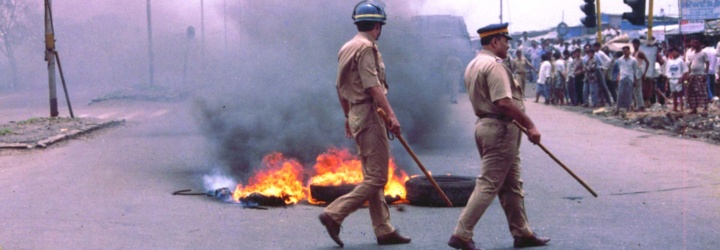 Two men in uniforms walk past tires burning in the middle of a road toward a crowd