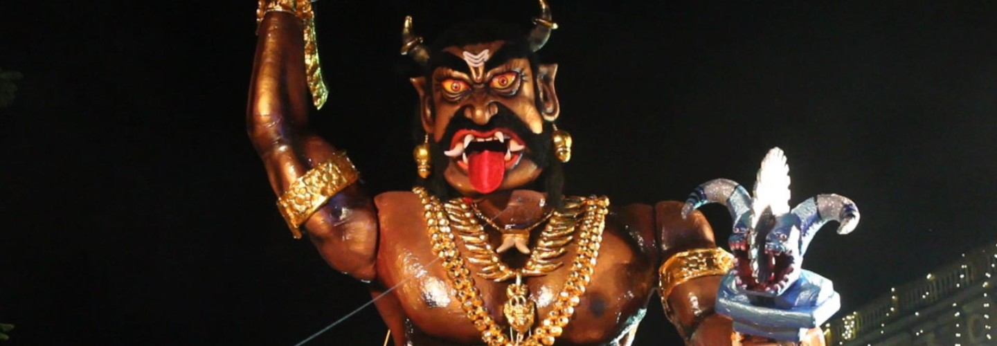 A large statue of a deity with tongue stuck out holding a staff before a black background