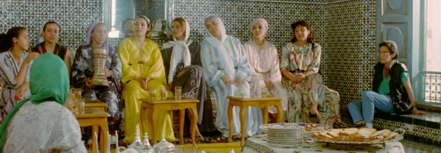 A group of women in colorful clothes seated along the patterned wall of a building