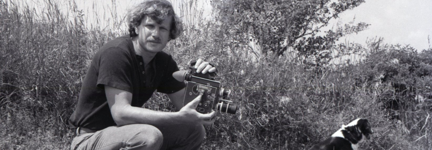 Black and white image of a man with light skin tone crouching in a field with a 16mm camera with a dog nearby