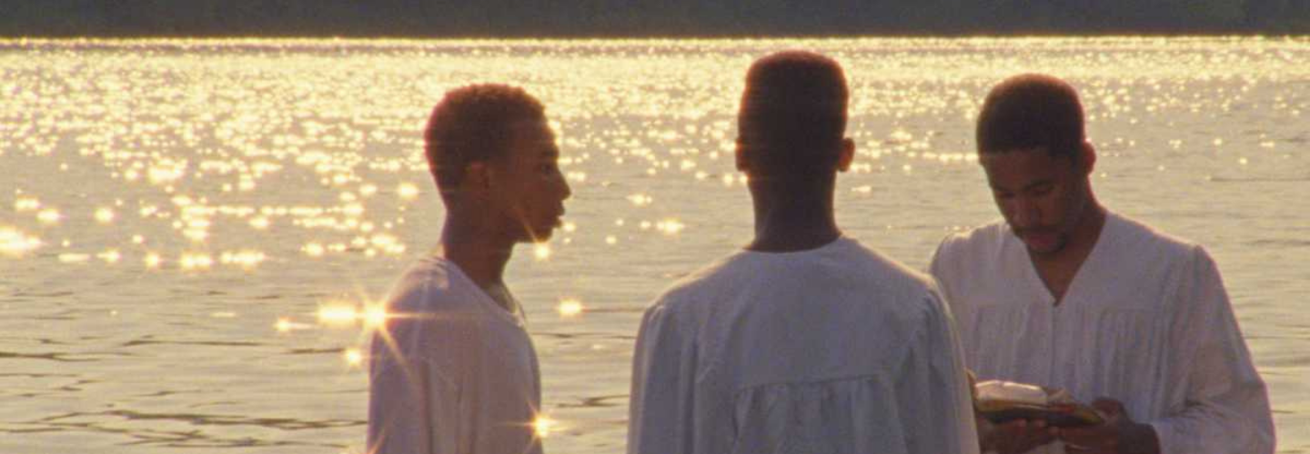 Three young Black men in white clothing stand in a river during the golden hour preparing for a baptism