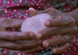 Light pink quartz stone held by hands with dark skin tone. Fuschia and blue printed cloth in the backdrop.