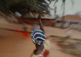 A person with a blue and white head wrap raises their arm surrounded by a blurry landscape 