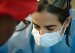 A woman with light skin and dark hair wearing a surgical mask and blue scrubs leans in toward another person with dark skin tone, glasses, and a red cap 