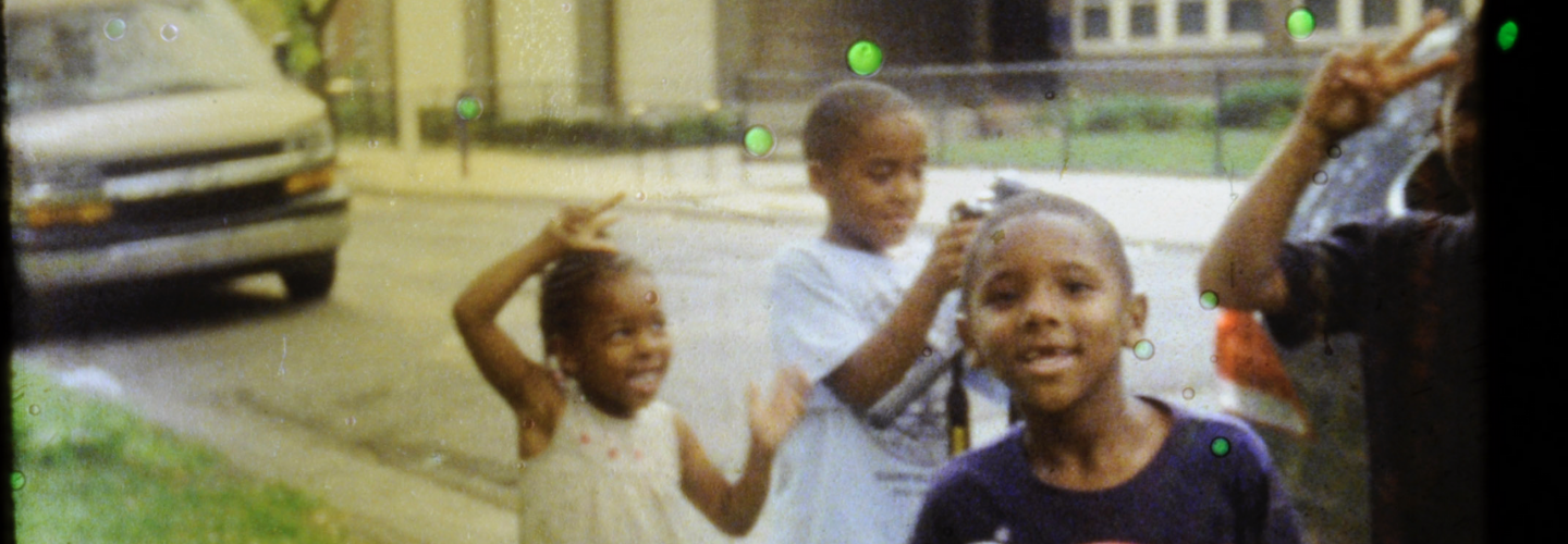 Four Black kids joyfully pose for a photo on a quiet street with parked cars