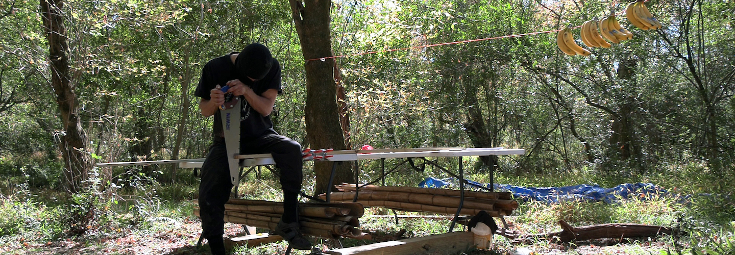 A figure in a balaclava and dark clothes standing in the forest uses a saw to cut a piece of wood.