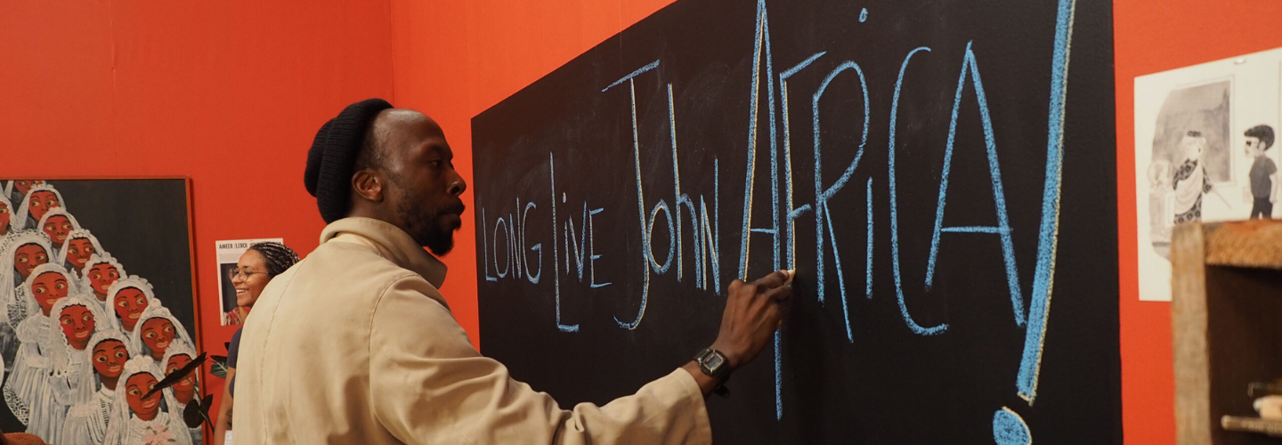 A Black man in a tan coat writes "Long Live John Africa!" on a blackboard in a red room