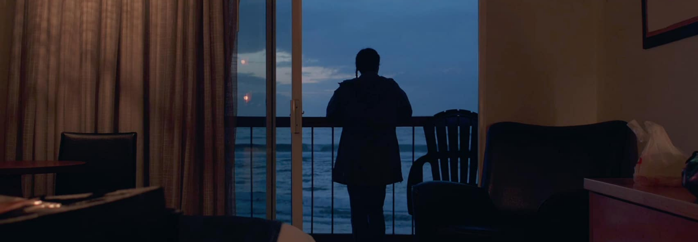 Facing away, a person stands on the balcony of a dimly lit bedroom that opens up to a view of the ocean 