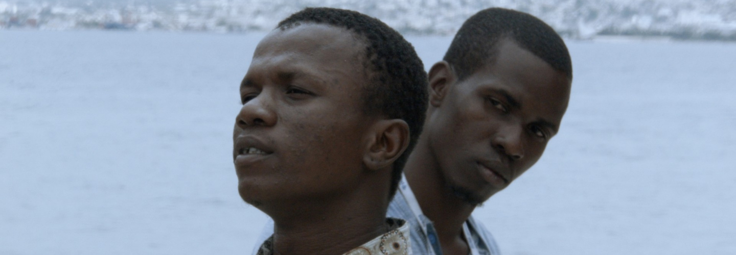 A close-up on two Black men stand beside one another looking away with the sea behind them