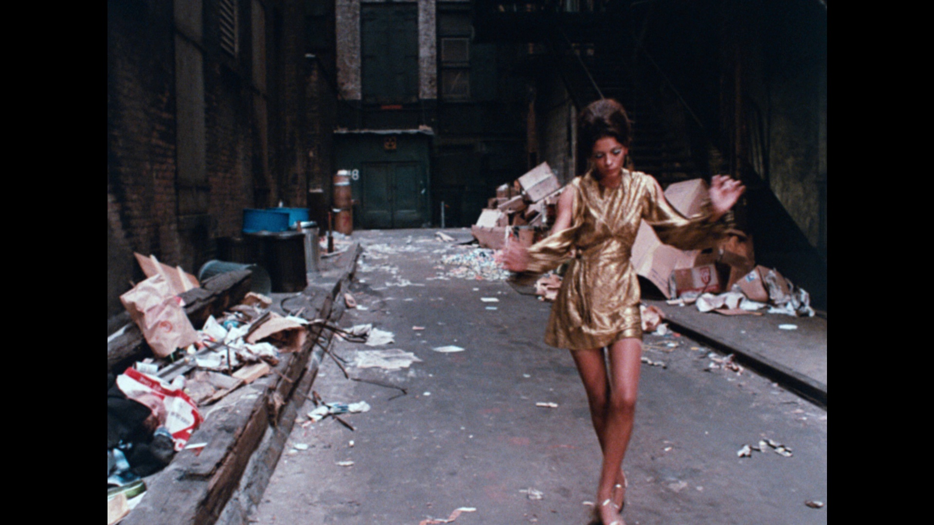 An African American woman wearing a short gold 1960s dress dances in an alley