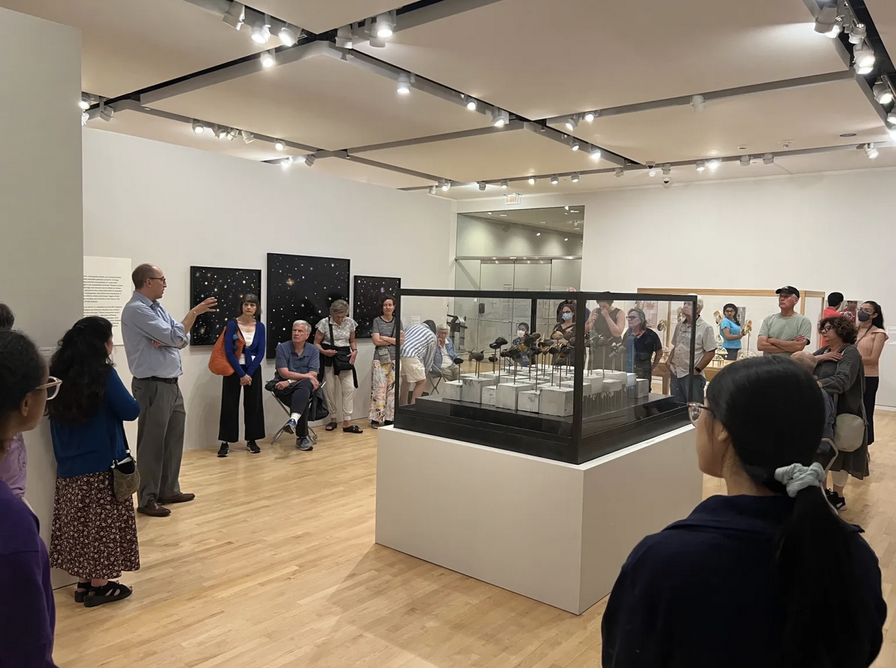 listeners stand around glass case in Block gallery as man in blue shirt speaks