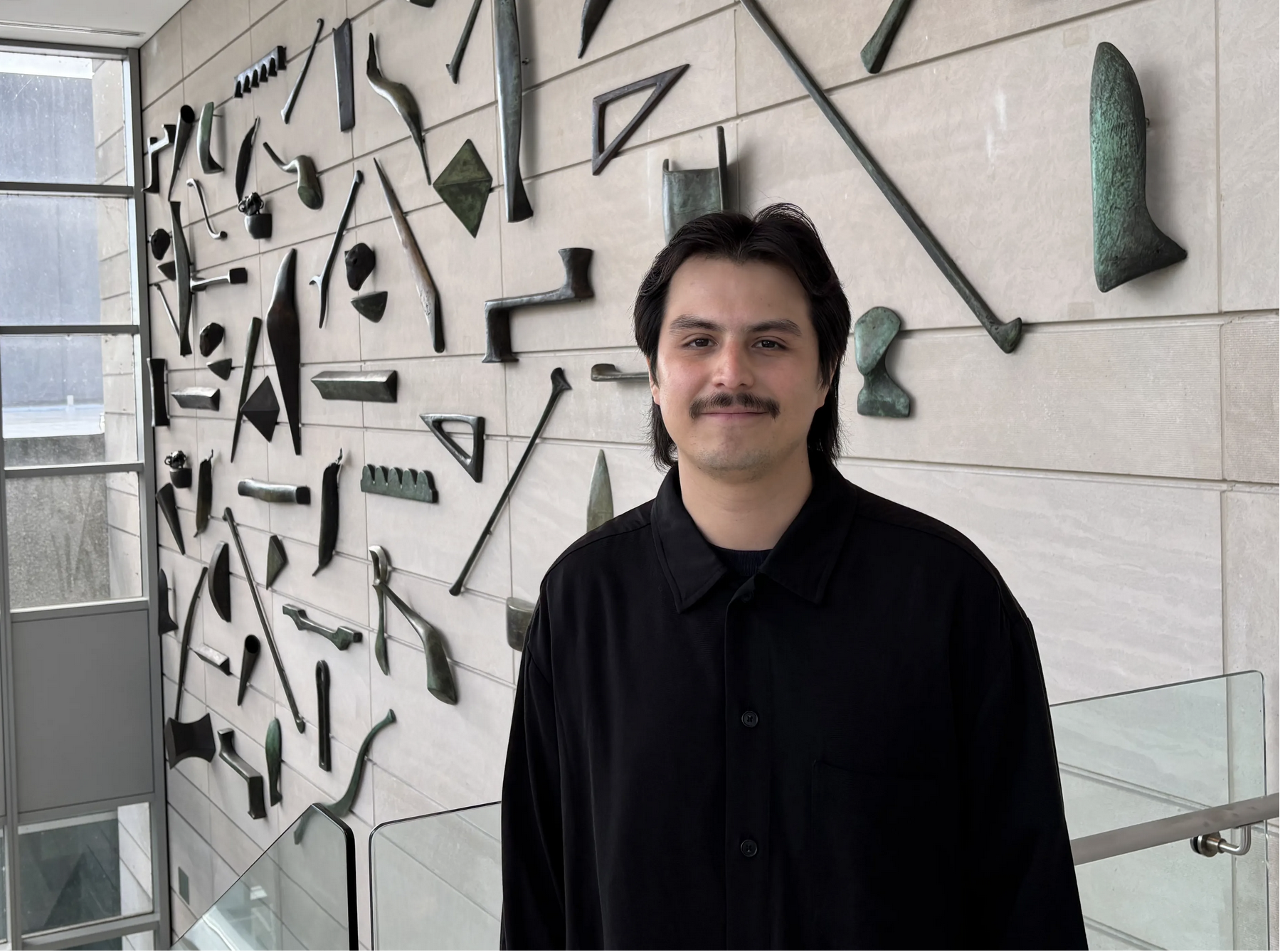 Man with dark hair and mustache stands in front of windows and sculpture-adorned wall