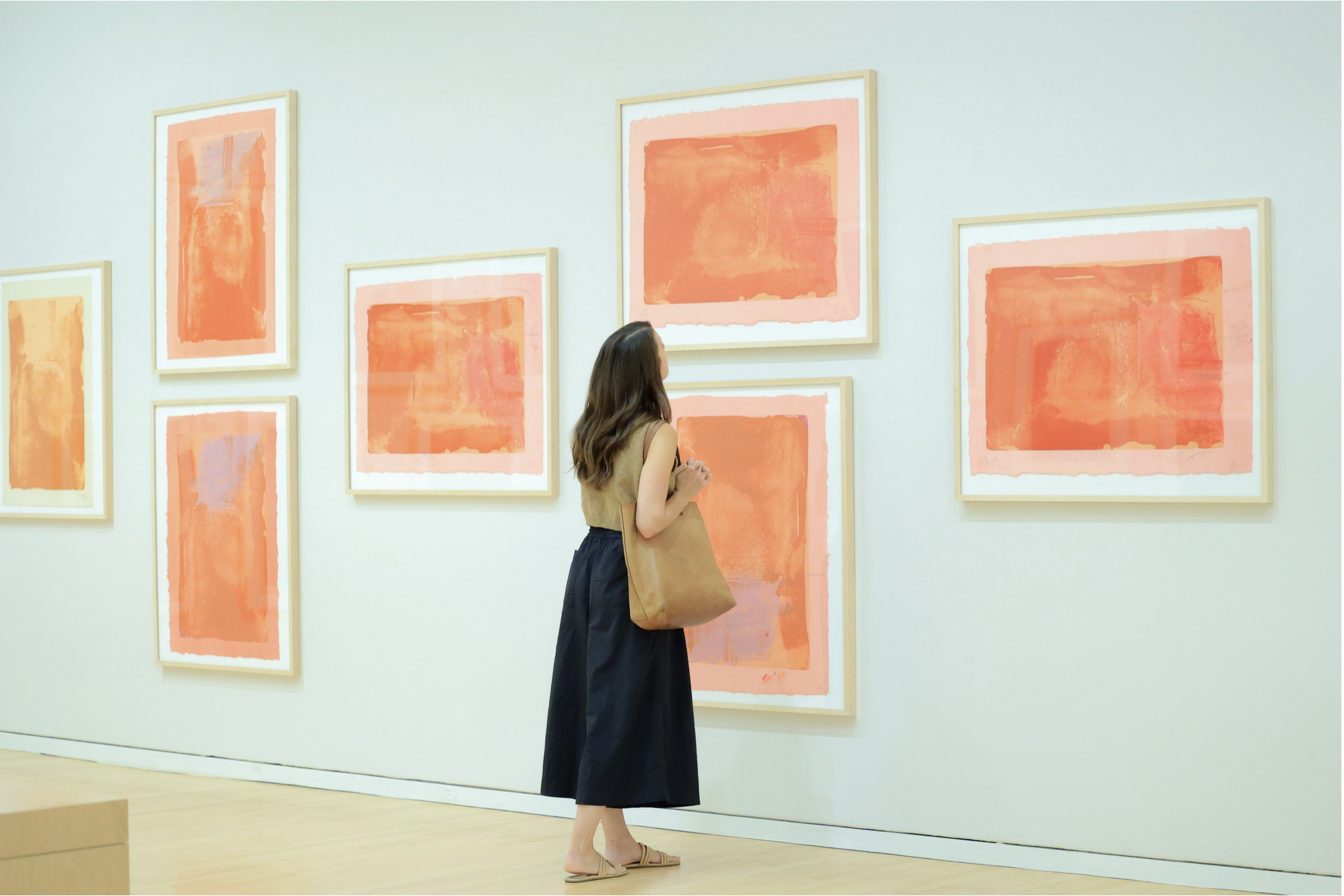 Brown-haired woman looks closely at one of seven similar orange prints on gallery wall