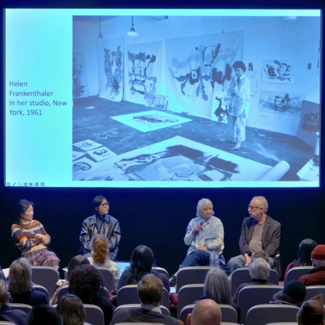 Panel of four speakers sits in front of auditorium screen displaying photo of Helen Frankenthaler in her studio