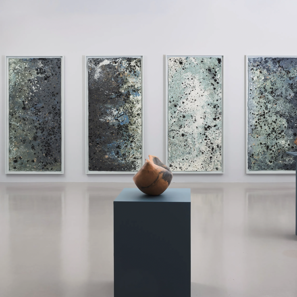 sculpture stands on pedestal in front of wall hangings in gallery space