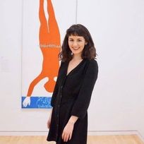 Person with shoulder-length, dark curly hair and front bangs. She wears a long-sleeved, black dress and stands in front of a painting of an up-side down, orange person.  